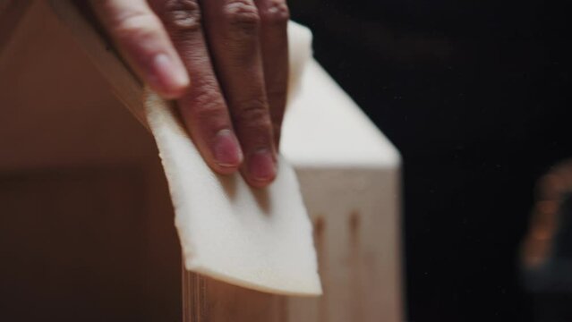 Unidentifiable man is sanding the edges of a wooden birdhouse with sandpaper. Close-up carpentry training, male hobby