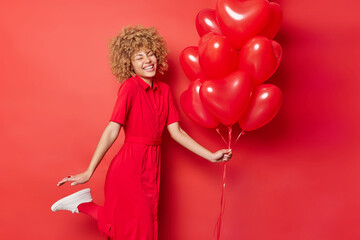 Happy energetic curly haired woman has fun dances and raises leg expresses positive emotions holds bunch of inflated heart shaped helium balloons comes on party isolated over vivid red background.