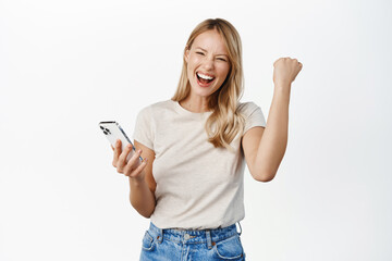 Enthusiastic young woman winning on mobile phone, rejoicing, using smartphone app, celebrating, triumphing on cellphone, white background - 488423108
