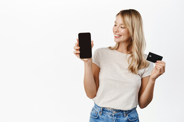 Beautiful girl smiling, showing smartphone screen and credit card, paying, shopping application app interface, standing over white background