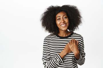 Image of cute african woman looking with love, tenderness and care, holding hands near heart, appreciate smth, standing over white background