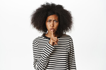 Image of Black female model shows shush, quiet gesture, taboo sign, press fingers to lips, tell secret, making hush sign, standing over white background