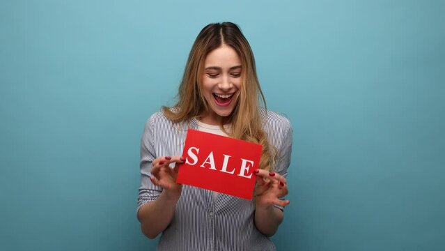 Portrait of beautiful young wavy haired woman holding sale word, looking smiling at camera, presenting discounts, wearing striped shirt. Indoor studio shot isolated on blue background.
