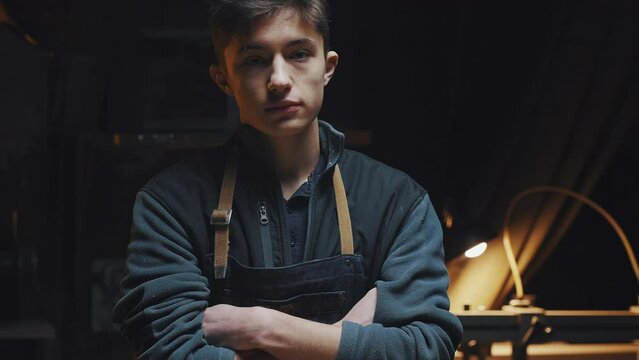 Young man in a working apron stands near the desktop on which carpentry tools and wooden blanks lie. Portrait of a master in the workshop, behind him stands a lathe, a woodworking machine