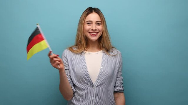 Portrait of pleasant looking woman with wavy hair holding Germany flag, celebrating Day of Germany - 3th October, wearing striped shirt. Indoor studio shot isolated on blue background.