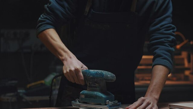Close portrait of a carpenter with a grinder in his hands. A young apprentice guy works in a carpentry workshop, polishing parts for handmade birdhouses