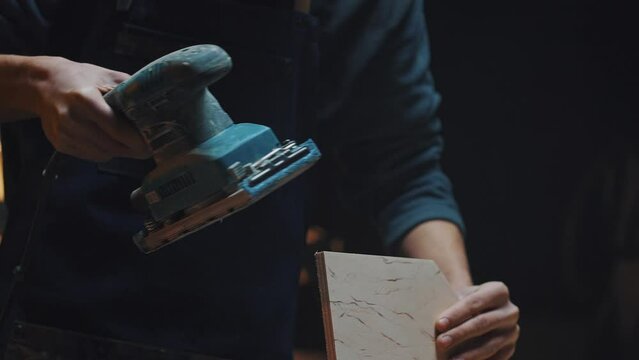 Side view of a carpenters hands with a grinder. A young apprentice guy works in a carpentry workshop, polishing parts for handmade birdhouses and wooden houses. Family workshop and hobby concept