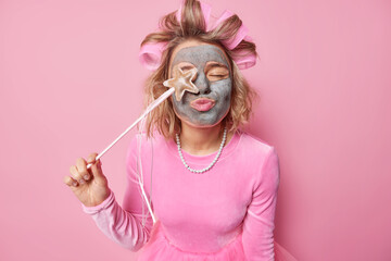 Horizontal shot of female model keeps lips rounded covers eye with magic wand applies nourishing clay mask to reduce fine lines makes hairstyle wears festive dress and necklace isolated on pink wall