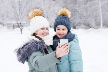 Fototapeta na wymiar Two smiling girls in warm clothes make a video call on a smartphone in a snowy winter park. Lifestyle use of technology
