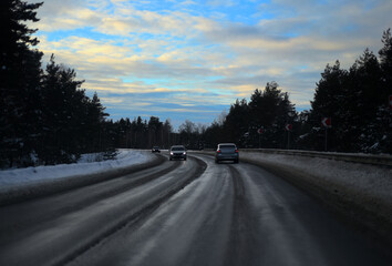 Winter. Cars drive along the road with a turn through the forest. There are snow-covered firs along the road, a beautiful blue sky with milky clouds.