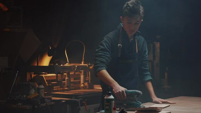 Cinematic video of a carpenter at work in a carpentry workshop. The guy polishes wooden parts with a grinder. Dark key, dust and small wood shavings are visible in the air