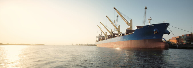 Large cargo ship (bulk Carrier, 187 meters length) loading in port terminal, cranes in the background. Freight transportation, logistics, global communications, economy, business, industry © Alex Stemmer