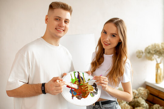 Portrait of happy smiling young pretty couple stand closely, posing, holding watercolor palette and brushes. Art therapy