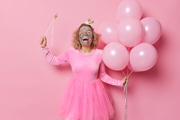 Positive woman prepares for birthday celebration applies beauty mask on face to look beautiful holds magic wand and bunch of helium balloons wears dress poses against pink background. Holidays