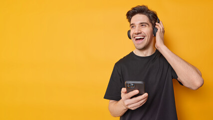 Horizontal shot of happy adult man dressed in casual black t shirt listens music via headphones...