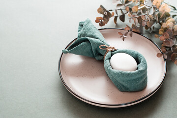 Obraz na płótnie Canvas A white chicken egg is wrapped in a napkin in the form of a rabbit on a plate and a branch of eucalyptus on a green background. Eco friendly happy easter.