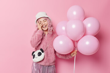Positive woman with pink hair keeps hand on face smiles gladfully dressed in outerwear has fun on special occasion enjoys celebration holds bunch of inflated balloons isolated over pink wall