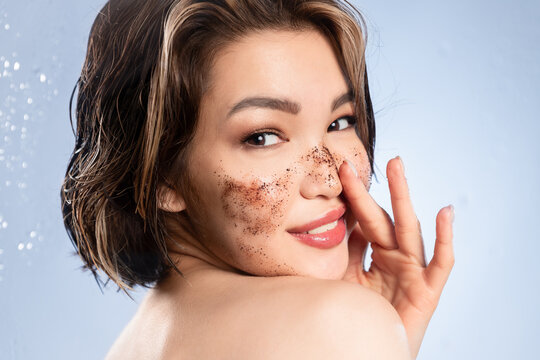 Playful young asian woman with bare shoulder scrubbing facial skin, cleansing face by cosmetic scrub, removing makeup for glowing pure healthy skin. Skincare daily routine.