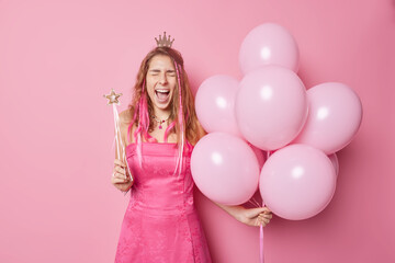 Obraz na płótnie Canvas Horizontal shot of emotive young woman exclaims loudly reacts on something holds inflated balloons and magic wand entertains children on party isolated over pink background. Celebration concept