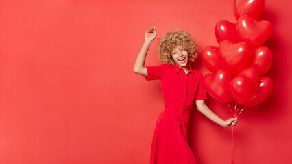 Energetic woman with blonde curly hair wears dress dances carefree with bunch of heart shaped balloons has fun celebrates holiday isolated over red background with copy space for promo. Valentines Day