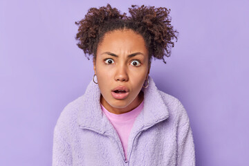Stunned indignant curly woman stares bugged eyes keeps mouth opened reacts on something unexpected and unpleasant dressed in warm fur jacket isolated over purple background. Oh no it cant be so