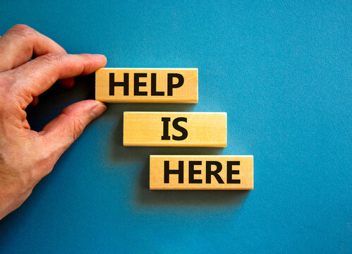Help is here and support symbol. Concept words Help is here on wooden blocks on beautiful blue table blue background. Businessman hand. Business, support, help is here concept. Copy space.