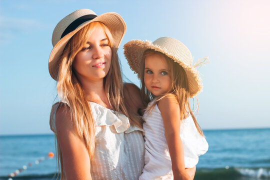 Happy mother and child walk along the sea, in hats, sundresses at sunset