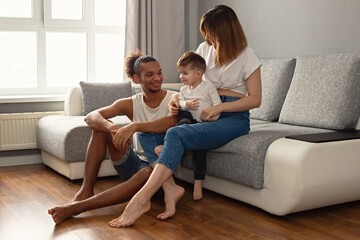 Happy, modern, multiracial family with a little boy in cozy home clothes sit on a gray sofa near a large window, smiling