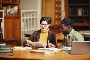 Fototapeta na wymiar Warm toned portrait of smiling adult woman studying in college library with African-American friend