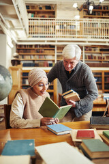 Vertical portrait senior professor talking to ethnic student wearing head cover in college library