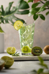 A green refreshing beverage with kiwi, lime and ice cubes in tall long glass with splashes on marble board in green plants setting
