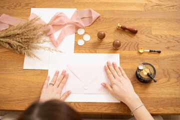 Cropped photo of woman hands making greeting envelope craft, decorate with pink ribbon, stamping. Top. Cereals on desk. 