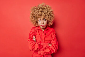 Displeased offended youg woman keeps arms folded looks unhappily away has hurt feelings has leaked makeup wears shirt isolated over red background. Moody gloomy female model feels depressed.