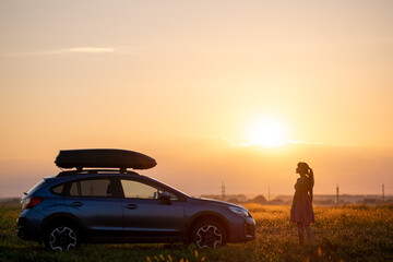 Fototapeta na wymiar Silhouette of female driver standing near her car on grassy field enjoying view of bright sunset. Young woman relaxing during road trip beside SUV vehicle