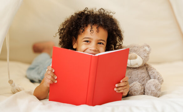 Cute little african american kid curly boy holding book lying in play tent with teddy bear toy