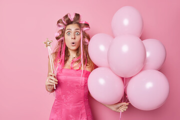 Obraz na płótnie Canvas Shocked stunned European woman makes hairstyle with rollers prepares for party wearrs dress holds magic wand inflated helium balloons isolated over pink background. Special occasion concept.