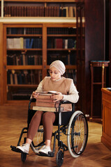 Full length portrait of young woman in wheelchair holding stack of books at college library