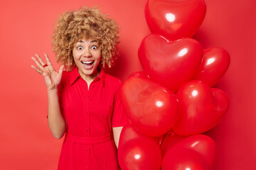 Surprised cheerful curly haired woman gets uexpected surprise gify on Valentines Day holds bunch of heart shaped balloons wears dress isolated over red background. People and celebration concept