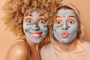 Close up shot of two young women make grimace at camera apply clay masks for skin treatment enjoy beauty procedures isolated over brown background. Pampering facial care and rejuvenation concept
