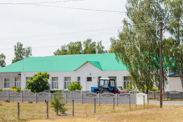 Fototapeta na wymiar Defocus blue tractor riding away in road on tree nature background. White building with green roof. Countryside area, school. Out of focus