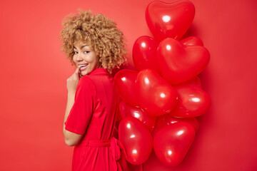 Happy young woman has romantic intrigued expression holds bunch of inflated heart balloons behind back wears dress isolated over vivid red background. People celebration and holidays concept