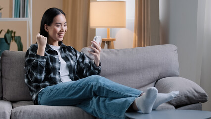 Asian girl woman sitting on sofa relaxing at home on couch playing mobile game wins getting new job...