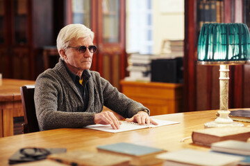 Portrait of senior man with vision impairment reading book in braille at table in classic library