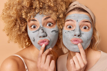 Close up shot of two shocked women stare impressed keep index fingers near folded lips apply nourishing clay mask to remove fine lines and pores stand closely to each other. Beauty time concept