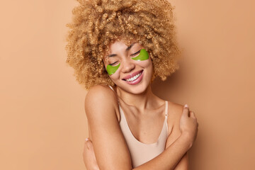 Tender cheerful European woman with curly bushy hair embraces own body wears t shirt smiles gently applies hydrogel green patches under eyes isolated over brown background. Wellness concept.
