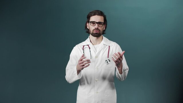 Young medical doctor in glasses and with statoscope wearing white coat in a green room talks about covid