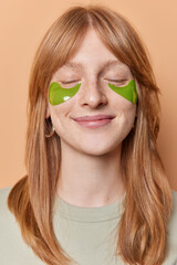 Vertical shot of redhead European girl keeps eyes closed smiles pleasantly enjoys softness of skin as undergoes beauty procedures regularly applies green hydrogel patches under eyes poses indoor.