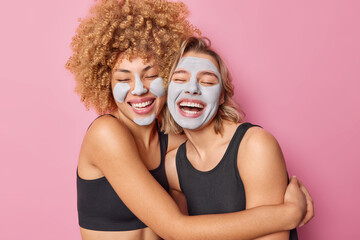 Horizontal shot of happy friendly young women embrace and have fun laugh gladfully keep eyes closed dressed in casual black t shirts wear beauty clay masks on faces isolated over pink background