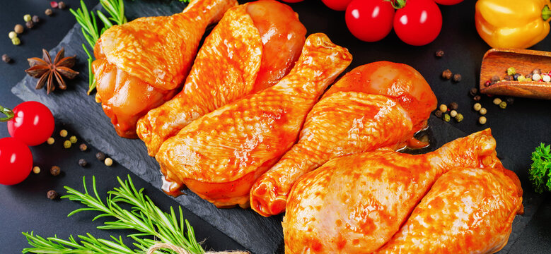 Marinated product, quick cooking.Barbecue marinated chicken dramstik on a black cooking board, with fresh rosemary leaves and fresh cherry tomatoes and various spices, close-up.Semi-finished product.