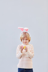 Obraz na płótnie Canvas Vertical waist up portrait of cute blonde boy wearing bunny ears and playing with Easter eggs against neutral white background, copy space